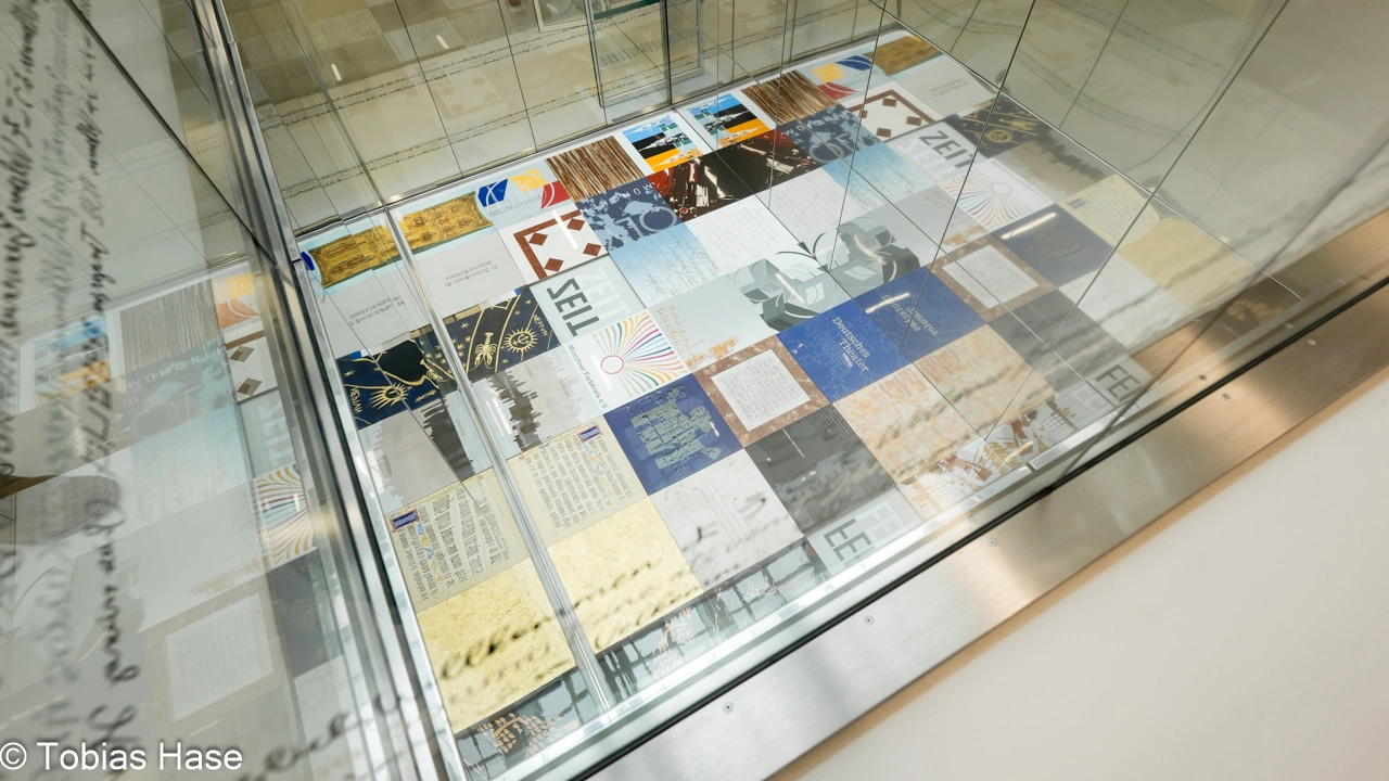 kreative-muster-fliesendesign-ausstellung.jpg Blick durch eine Glasscheibe auf kunstvoll gestaltete, quadratische Fliesen und Farbmuster, die am Boden ausgestellt sind.